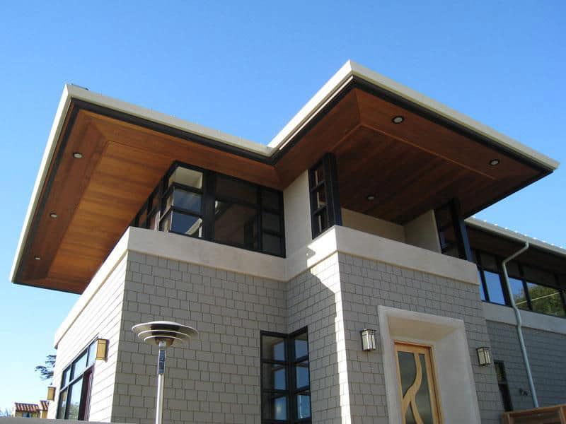 A modern home, featuring staggered grey brick stones, a glass and wood door, a balcony, and large windows. The eaves of the roof have recessed can lighting.
