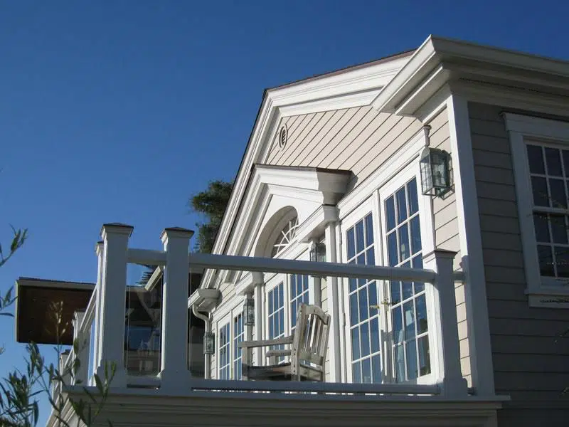 A colonial style home with a balcony with white bannisters and glass panels as the walls. The house has off white siding with white trim, and three sets of double French doors that access the balcony.