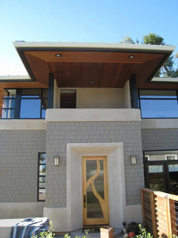 A modern home, featuring staggered grey brick stones, a glass and wood door, a balcony, and large windows. The eaves of the roof have recessed can lighting.