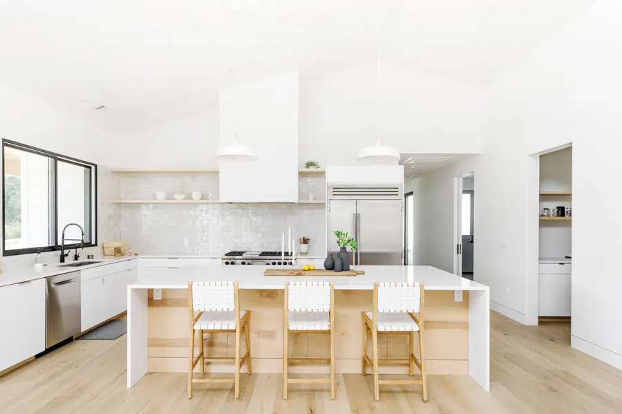 A farmhouse style home with a modern kitchen. There is a white center island with natural wood cabinets, and three chairs with white woven seats and natural wood legs.