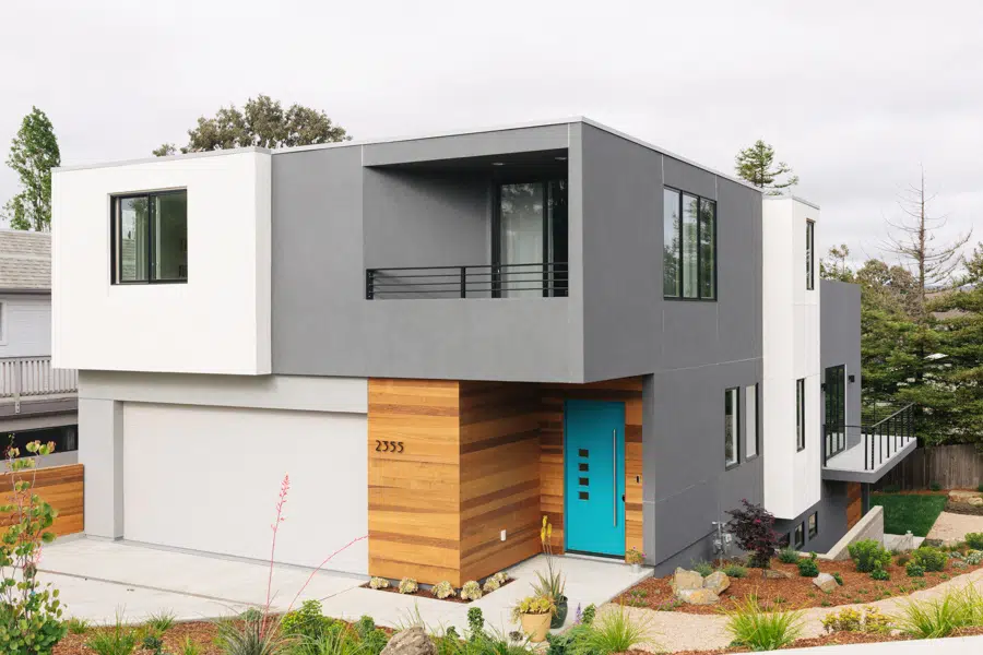 An exterior photo of a modern home with angular features with white and grey walls, accented with a wood entry way and a blue front door.