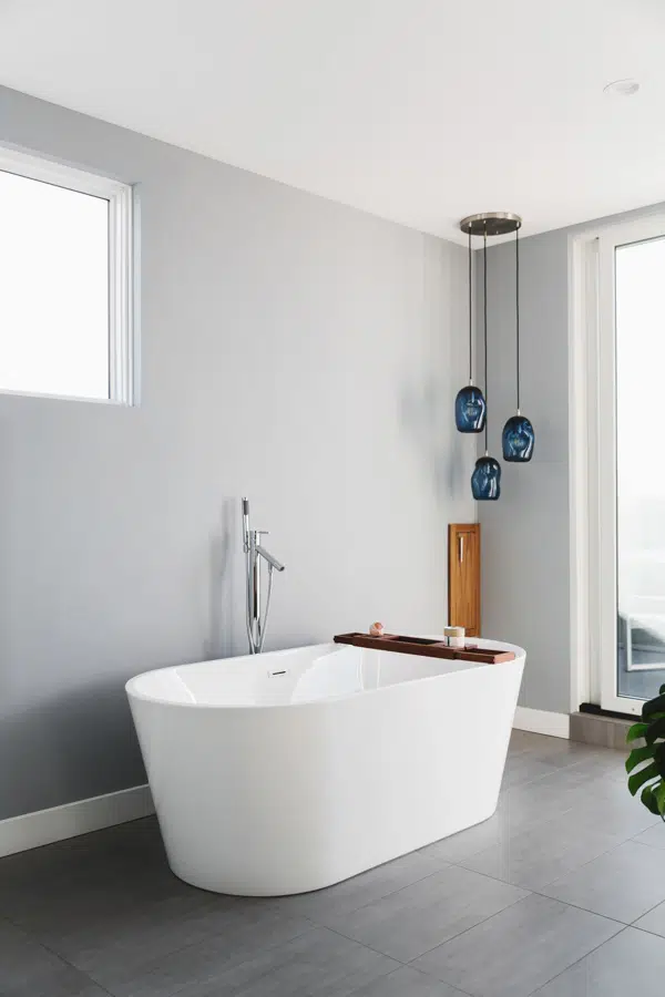 A bathroom in a modern style home, with grey walls and white baseboards, and a sleek white bathtub. There is a three-piece pendant light with blue glass shades in the corner.