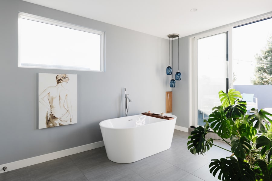 A bathroom in a modern style home, with grey walls and white baseboards, and a sleek white bathtub. There is a three-piece pendant light with blue glass shades in the corner.