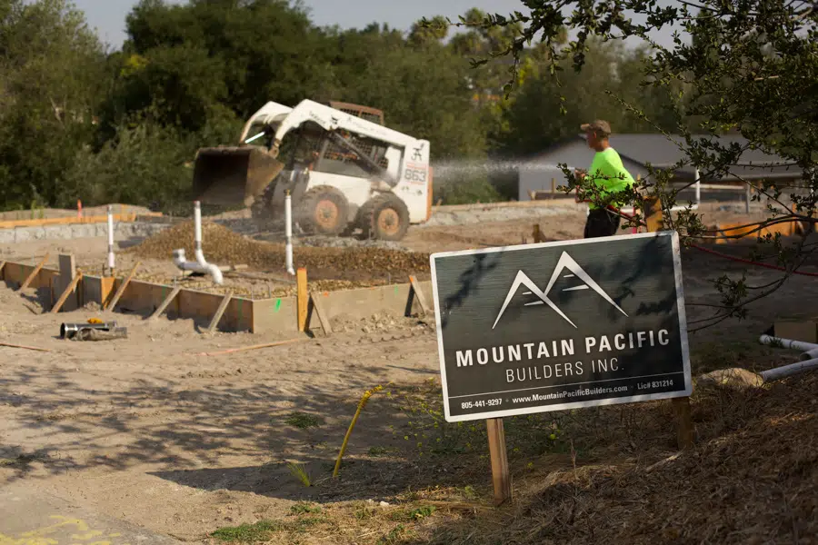 A construction site, with a white Bobcat 863 skid steer loader and a man in a yellow shirt spraying a water hose. There is a black and white sign with the Mountain Pacific Builders Inc. logo and information in the foreground.