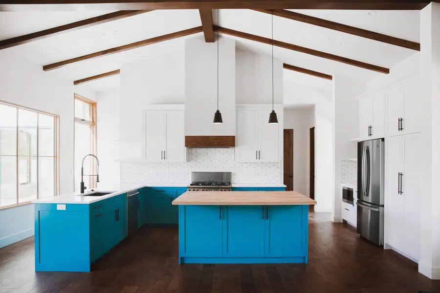 A Spanish Canyon style kitchen with a white ceiling, white walls, dark wood beams, and large windows. The cabinets are blue, with white countertops and a butcher block countertop on the island.
